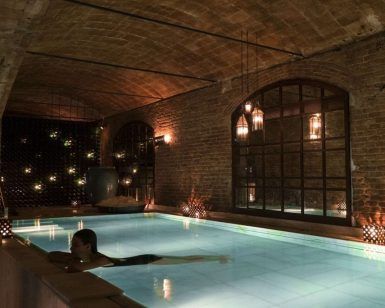 En una zona de piscina subterránea poco iluminada, con paredes de ladrillo y luces colgantes, que recuerda a la "Experiencia Baño termal con masaje en Aires de Barcelona", experimente momentos serenos de relajación. - ForTwo - Planes para parejas