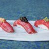 Tres piezas de sushi con diversas guarniciones se disponen elegantemente en un plato blanco sobre un fondo azul, evocando las vibrantes experiencias culinarias de la Experiencia Kaitai en Tunateca Balfegó de Barcelona. - ForTwo - Planes para parejas