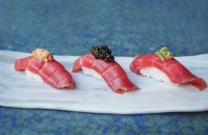 Tres piezas de sushi con diversas guarniciones se disponen elegantemente en un plato blanco sobre un fondo azul, evocando las vibrantes experiencias culinarias de la Experiencia Kaitai en Tunateca Balfegó de Barcelona. - ForTwo - Planes para parejas