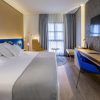 La moderna habitación del Hotel Barceló V Centenario (Cáceres) ofrece una cama espaciosa, televisor, escritorio y silla. La luz natural entra por una ventana con cortinas transparentes, creando un ambiente acogedor. - ForTwo - Planes para parejas
