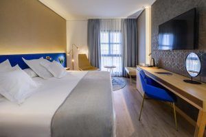 La moderna habitación del Hotel Barceló V Centenario (Cáceres) ofrece una cama espaciosa, televisor, escritorio y silla. La luz natural entra por una ventana con cortinas transparentes, creando un ambiente acogedor. - ForTwo - Planes para parejas