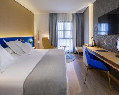 La moderna habitación del Hotel Barceló V Centenario (Cáceres) ofrece una cama espaciosa, televisor, escritorio y silla. La luz natural entra por una ventana con cortinas transparentes, creando un ambiente acogedor. - ForTwo - Planes para parejas