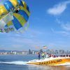 La embarcación amarilla con la etiqueta "Hacer parasailing en Barcelona" se desliza por el océano, remolcando un vibrante parasail frente al impresionante horizonte de Barcelona. - ForTwo - Planes para parejas