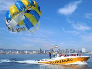 La embarcación amarilla con la etiqueta "Hacer parasailing en Barcelona" se desliza por el océano, remolcando un vibrante parasail frente al impresionante horizonte de Barcelona. - ForTwo - Planes para parejas