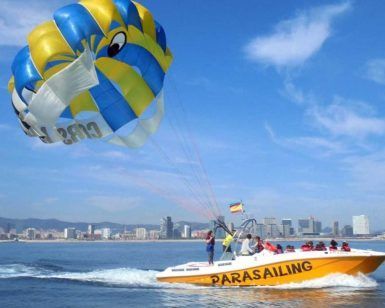 La embarcación amarilla con la etiqueta "Hacer parasailing en Barcelona" se desliza por el océano, remolcando un vibrante parasail frente al impresionante horizonte de Barcelona. - ForTwo - Planes para parejas