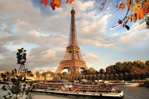 Contra un cielo nublado de París, "Descubre París en barco y bus y visita la Torre Eiffel" captura la majestuosa vista mientras los barcos se deslizan por el Sena enmarcados por hojas de otoño. - ForTwo - Planes para parejas