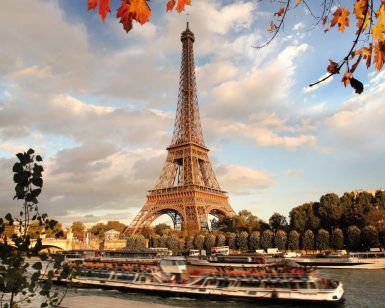 Contra un cielo nublado de París, "Descubre París en barco y bus y visita la Torre Eiffel" captura la majestuosa vista mientras los barcos se deslizan por el Sena enmarcados por hojas de otoño. - ForTwo - Planes para parejas