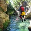 Alguien se lanza al río entre las rocas, sumergiéndose en la emoción del "Barranquismo en Granada - Tramo acuático en barranco de Lentegí", mientras otros nadan delante con cascos y equipos de protección. - ForTwo - Planes para parejas