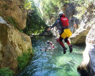 Alguien se lanza al río entre las rocas, sumergiéndose en la emoción del "Barranquismo en Granada - Tramo acuático en barranco de Lentegí", mientras otros nadan delante con cascos y equipos de protección. - ForTwo - Planes para parejas