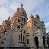 El recorrido "Descubre Montmartre y el Sacré Coeur (París)" captura la cúpula y las estatuas de la Basílica del Sacré-Coeur, bellamente iluminadas por el suave resplandor de un atardecer parisino contra un cielo nublado. - ForTwo - Planes para parejas