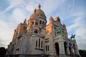 El recorrido "Descubre Montmartre y el Sacré Coeur (París)" captura la cúpula y las estatuas de la Basílica del Sacré-Coeur, bellamente iluminadas por el suave resplandor de un atardecer parisino contra un cielo nublado. - ForTwo - Planes para parejas