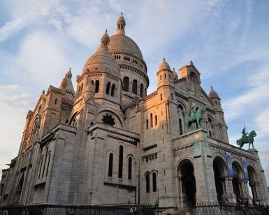 El recorrido "Descubre Montmartre y el Sacré Coeur (París)" captura la cúpula y las estatuas de la Basílica del Sacré-Coeur, bellamente iluminadas por el suave resplandor de un atardecer parisino contra un cielo nublado. - ForTwo - Planes para parejas