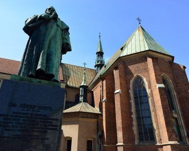 Ubicada en el histórico casco antiguo de Cracovia, una estatua adorna una clásica iglesia de ladrillo con ventanas arqueadas y techos verdes que brillan bajo un cielo azul claro, encarnando el encanto de este encantador barrio judío explorado durante la Visita guiada por el casco antiguo y el barrio judío (Cracovia). - ForTwo - Planes para parejas