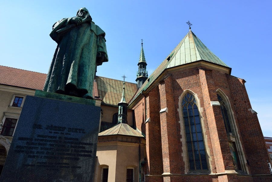 Ubicada en el histórico casco antiguo de Cracovia, una estatua adorna una clásica iglesia de ladrillo con ventanas arqueadas y techos verdes que brillan bajo un cielo azul claro, encarnando el encanto de este encantador barrio judío explorado durante la Visita guiada por el casco antiguo y el barrio judío (Cracovia). - ForTwo - Planes para parejas