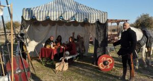 Dentro de una tienda, personas vestidas con trajes medievales disfrutan de la comida, evocando el pasado histórico de Toledo durante la Batalla medieval de arqueros; afuera, un hombre está listo con un escudo. - ForTwo - Planes para parejas
