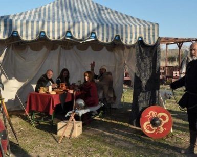 Dentro de una tienda, personas vestidas con trajes medievales disfrutan de la comida, evocando el pasado histórico de Toledo durante la Batalla medieval de arqueros; afuera, un hombre está listo con un escudo. - ForTwo - Planes para parejas