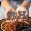 Primer plano de unas manos sosteniendo las letras metálicas B y H sobre una tarta en una mesa de madera, evocando el encanto artesanal de Bendita Helena (Barcelona). - ForTwo - Planes para parejas