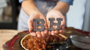 Primer plano de unas manos sosteniendo las letras metálicas B y H sobre una tarta en una mesa de madera, evocando el encanto artesanal de Bendita Helena (Barcelona). - ForTwo - Planes para parejas