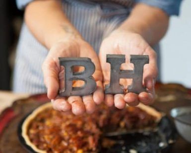 Primer plano de unas manos sosteniendo las letras metálicas B y H sobre una tarta en una mesa de madera, evocando el encanto artesanal de Bendita Helena (Barcelona). - ForTwo - Planes para parejas