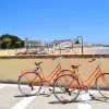 Dos bicicletas naranjas del "Tour guiado en Bicicleta por la ciudad (Cádiz)" están aparcadas junto a un muro de playa bajo un cielo despejado, con arena y bañistas al fondo. - ForTwo - Planes para parejas
