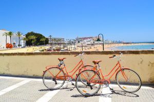 Dos bicicletas naranjas del "Tour guiado en Bicicleta por la ciudad (Cádiz)" están aparcadas junto a un muro de playa bajo un cielo despejado, con arena y bañistas al fondo. - ForTwo - Planes para parejas