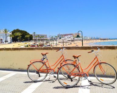Dos bicicletas naranjas del "Tour guiado en Bicicleta por la ciudad (Cádiz)" están aparcadas junto a un muro de playa bajo un cielo despejado, con arena y bañistas al fondo. - ForTwo - Planes para parejas