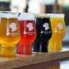 Descubre la Cata de cervezas en BlackLab en Barcelona: una bandeja de madera con cuatro coloridas cervezas artesanales, cada vaso con el logo de BlackLab. ¡Disfruta de una experiencia de sabor única!. - ForTwo - Planes para parejas