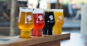 Descubre la Cata de cervezas en BlackLab en Barcelona: una bandeja de madera con cuatro coloridas cervezas artesanales, cada vaso con el logo de BlackLab. ¡Disfruta de una experiencia de sabor única!. - ForTwo - Planes para parejas