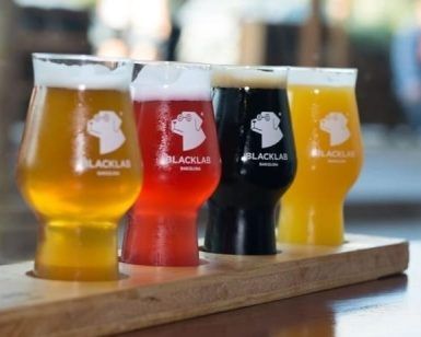 Descubre la Cata de cervezas en BlackLab en Barcelona: una bandeja de madera con cuatro coloridas cervezas artesanales, cada vaso con el logo de BlackLab. ¡Disfruta de una experiencia de sabor única!. - ForTwo - Planes para parejas