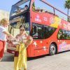 Dos mujeres vestidas de verano estudian un mapa junto a un autobús rojo de dos pisos con "Billetes para el Bus turístico de Córdoba" en la mano, bajo una calle soleada y bordeada de palmeras que recuerda a Córdoba. - ForTwo - Planes para parejas