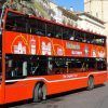 Un radiante cartel rojo de "Entradas para el bus turístico de Valencia" se desliza por la ciudad, con un impresionante telón de fondo de edificios históricos en un día soleado. - ForTwo - Planes para parejas