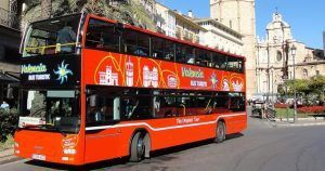 Un radiante cartel rojo de "Entradas para el bus turístico de Valencia" se desliza por la ciudad, con un impresionante telón de fondo de edificios históricos en un día soleado. - ForTwo - Planes para parejas