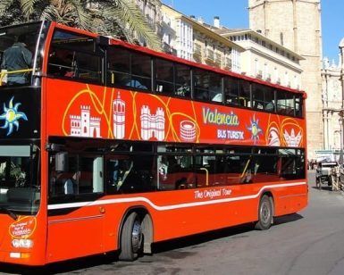 Un radiante cartel rojo de "Entradas para el bus turístico de Valencia" se desliza por la ciudad, con un impresionante telón de fondo de edificios históricos en un día soleado. - ForTwo - Planes para parejas