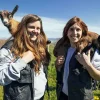 Dos mujeres disfrutan de la experiencia, sonriendo con cabras sobre sus hombros frente a los exuberantes campos y cielos de Muntanyola durante la "Visita las cabras de Peyu con cata incluida" en Barcelona. - ForTwo - Planes para parejas