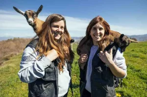Dos mujeres disfrutan de la experiencia, sonriendo con cabras sobre sus hombros frente a los exuberantes campos y cielos de Muntanyola durante la "Visita las cabras de Peyu con cata incluida" en Barcelona. - ForTwo - Planes para parejas