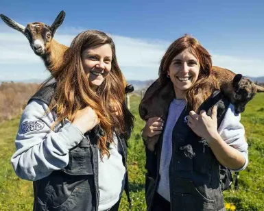 Dos mujeres disfrutan de la experiencia, sonriendo con cabras sobre sus hombros frente a los exuberantes campos y cielos de Muntanyola durante la "Visita las cabras de Peyu con cata incluida" en Barcelona. - ForTwo - Planes para parejas
