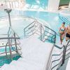 Una pareja en traje de baño sube las escaleras de una luminosa piscina cubierta en Caldea Spa + Comida o cena para dos (Andorra), que muestra un diseño moderno y una fuente de agua circular, capturando el ambiente sereno de Andorra. - ForTwo - Planes para parejas