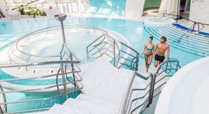 Una pareja en traje de baño sube las escaleras de una luminosa piscina cubierta en Caldea Spa + Comida o cena para dos (Andorra), que muestra un diseño moderno y una fuente de agua circular, capturando el ambiente sereno de Andorra. - ForTwo - Planes para parejas