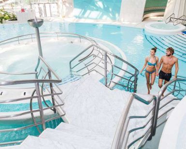 Una pareja en traje de baño sube las escaleras de una luminosa piscina cubierta en Caldea Spa + Comida o cena para dos (Andorra), que muestra un diseño moderno y una fuente de agua circular, capturando el ambiente sereno de Andorra. - ForTwo - Planes para parejas