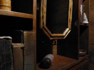 Estantes de madera tenuemente iluminados con libros antiguos, una botella inclinada y un espejo con marco de madera que refleja la oscuridad evocan el ambiente misterioso de Capitan Pessoa - Un juego para 2 de Retobox Escape Room (Barcelona). - ForTwo - Planes para parejas