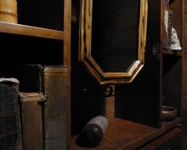 Estantes de madera tenuemente iluminados con libros antiguos, una botella inclinada y un espejo con marco de madera que refleja la oscuridad evocan el ambiente misterioso de Capitan Pessoa - Un juego para 2 de Retobox Escape Room (Barcelona). - ForTwo - Planes para parejas
