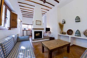 Relájese en una acogedora habitación en "Escapada en casa rural en el Parque Nacional de Sierra Nevada (Granada)", con sofás de cuadros azules, una mesa de madera y una chimenea. Disfrute de una decoración rústica con paredes blancas que evoca la encantadora sencillez granadina. - ForTwo - Planes para parejas