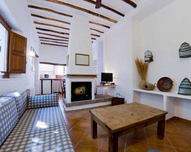 Relájese en una acogedora habitación en "Escapada en casa rural en el Parque Nacional de Sierra Nevada (Granada)", con sofás de cuadros azules, una mesa de madera y una chimenea. Disfrute de una decoración rústica con paredes blancas que evoca la encantadora sencillez granadina. - ForTwo - Planes para parejas