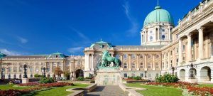 Explora la belleza arquitectónica del Castillo de Buda en Budapest, con cúpulas verdes, una fachada ornamentada y una estatua de jardín bajo un cielo azul claro con el "Tour por el castillo de Buda (Budapest). - ForTwo - Planes para parejas