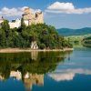 Un castillo medieval en una colina junto al lago, en medio de una exuberante vegetación, se refleja en el agua tranquila bajo un cielo azul, que recuerda la experiencia que ofrece "Descenso en barco al río Dunajec y visita al castillo de Niedzica (Cracovia). - ForTwo - Planes para parejas
