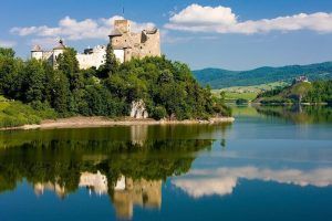 Un castillo medieval en una colina junto al lago, en medio de una exuberante vegetación, se refleja en el agua tranquila bajo un cielo azul, que recuerda la experiencia que ofrece "Descenso en barco al río Dunajec y visita al castillo de Niedzica (Cracovia). - ForTwo - Planes para parejas
