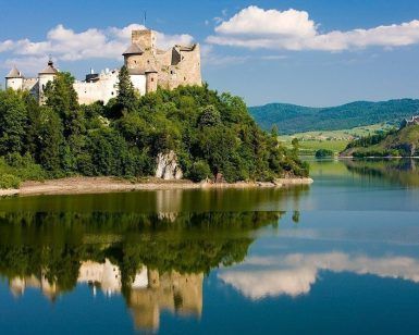 Un castillo medieval en una colina junto al lago, en medio de una exuberante vegetación, se refleja en el agua tranquila bajo un cielo azul, que recuerda la experiencia que ofrece "Descenso en barco al río Dunajec y visita al castillo de Niedzica (Cracovia). - ForTwo - Planes para parejas
