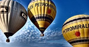 Tres globos aerostáticos con la marca Globus Kon Tiki flotan con gracia bajo un cielo azul salpicado de nubes, ofreciendo una inolvidable "Oferta de vuelo en globo en Cataluña con Globus Kon Tiki" sobre los paisajes escénicos de Cataluña. - ForTwo - Planes para parejas