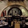 Disfrute de un ambiente gastronómico medieval en el Palau Requesens de Barcelona, con paredes de piedra rústica, mesas redondas y puertas arqueadas con la experiencia "Cena a lo medieval con espectáculo". - ForTwo - Planes para parejas