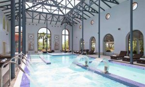 La piscina cubierta del Circuito Yhi Spa Meliá Villaitana (Alicante) cuenta con tumbonas, ventanas arqueadas con vigas de acero y elementos de agua en cascada, creando un ambiente sereno en este lujoso oasis. - ForTwo - Planes para parejas
